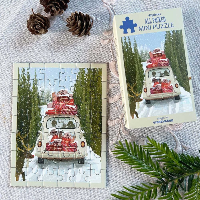 The All Packed | 42 Piece Jigsaw Puzzle by ViSSEVASSE, shown completed alongside its box, features a white car carrying red luggage as it drives through snowy pine trees. Pinecones and greenery are arranged nearby on a textured fabric surface.