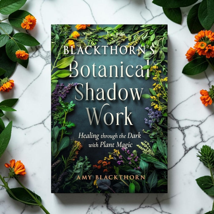 A book titled 'Blackthorn's Botanical Shadow Work' by Amy Blackthorn is displayed flat on a white marble surface, surrounded by green leaves and orange flowers. The book cover features a dark background decorated with botanical illustrations and white text.