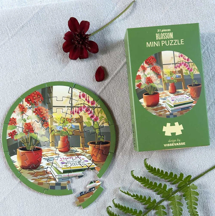 A vibrant, round 31-piece jigsaw puzzle from ViSSEVASSE, featuring a lively plant scene, rests beside its โBlossom | 31 Piece Jigsaw Puzzleโ box on a white surface, accompanied by a red flower and fern leafโmaking this compact travel puzzle ideal for puzzlers on the move.