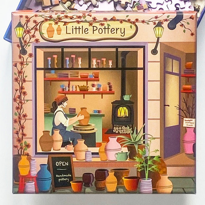 ViSSEVASSEβs βLittle Pottery | 150 Piece Jigsaw Puzzleβ features a charming pottery shop named Little Pottery, complete with shelves of handmade ceramics, an artist shaping clay at the wheel, lush plants outside, and an OPEN chalkboard sign by the sidewalkβideal for anyone seeking inspiration or wishing to assemble their own unique scene piece by piece.