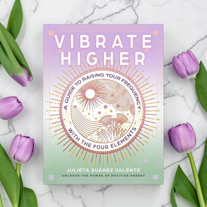 Book cover for 'Vibrate Higher' by Julieta Suárez Valente, styled in a flat-lay on a white marble surface with purple tulips. The cover features a pastel gradient transitioning from lavender to mint green, with a central circular illustration of suns, stars, and elements in gold and pink.