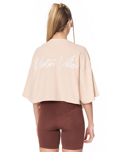 A woman shown from the back wearing a light beige cropped t-shirt with 'Martin Valen' written in white script across the back, paired with brown bike shorts, against a plain white background.