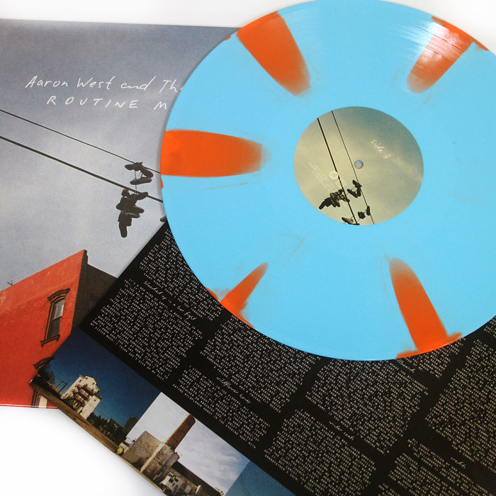 A blue vinyl with orange splashes rests on an album cover featuring Aaron West and the Roaring Twenties’ birds perched on power lines by a red building, with a black inner sleeve showing white Pinwheel Tracklisting text.