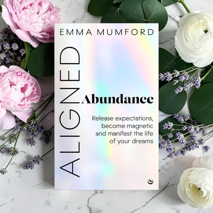 Book cover for 'Aligned Abundance' by Emma Mumford, styled in a flat lay with pink and white flowers and green leaves on a white marble background.