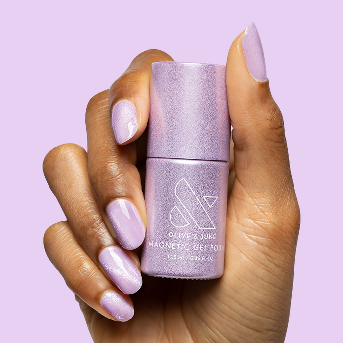 Hand with nails painted in Amethyst Velvet - Gel pastel lavender magnetic gel polish holding matching bottle against light purple background