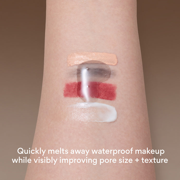 Close-up of a person's forearm with four makeup swatches applied: a beige cream, mascara smudge, a red lipstick layer, and a white cream. A translucent clear product is being pressed over the center, with text at the bottom reading 'Quickly melts away waterproof makeup while visibly improving pore size + texture'.