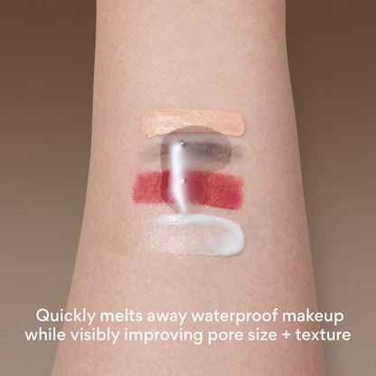 Close-up of a person's forearm with four makeup swatches applied: a beige cream, mascara smudge, a red lipstick layer, and a white cream. A translucent clear product is being pressed over the center, with text at the bottom reading 'Quickly melts away waterproof makeup while visibly improving pore size + texture'.
