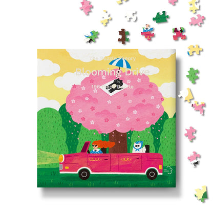 Blooming Drive | 196 Piece Jigsaw Puzzle by The Puzzle Theory features an illustration of a pink car, a person, and a cat beneath a blooming cherry tree, with scattered pieces around the box. Made from recycled paperboard, this charming springtime puzzle offers eco-friendly fun.