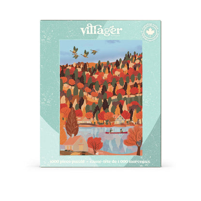 A Villager Puzzles “Changing Seasons” 1,000-piece jigsaw puzzle box featuring artwork by Marie-Laure Plano, depicting autumn trees, charming houses, a lake with two people canoeing, and birds in flight.