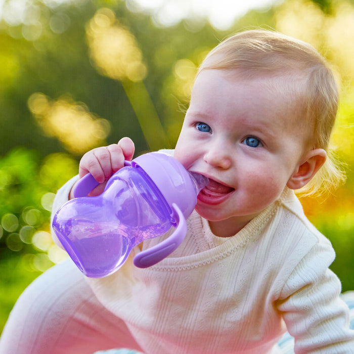 A baby with light hair and blue eyes smiles while holding a Clik-It 2-Handle Sippy Cup (2 Pack) | Purple / Aqua outdoors in soft sunlight.