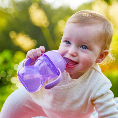 A baby with light hair and blue eyes smiles while holding a Clik-It 2-Handle Sippy Cup (2 Pack) | Purple / Aqua outdoors in soft sunlight.