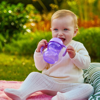 A baby in a white outfit sits on a blanket outdoors, smiling with greenery behind, while happily drinking from the Clik-It 2-Handle Sippy Cup (2 Pack) | Purple / Aqua.