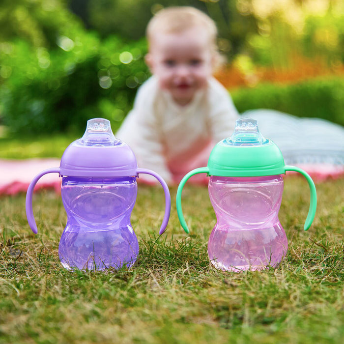 A baby on a blanket is blurred in the background, while two Clik-It 2-Handle Sippy Cup (2 Pack) | Purple / Aqua sit on the grass in the foreground.