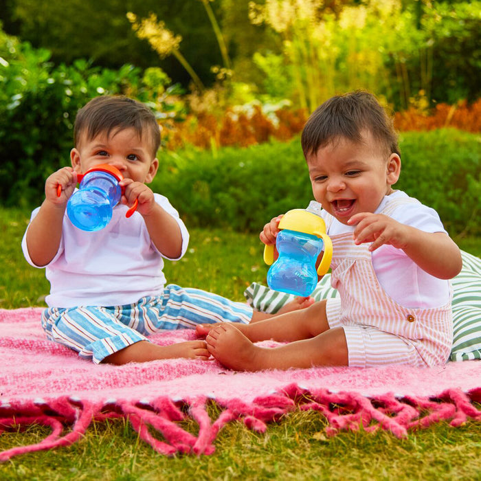 Two toddlers enjoy a sunny picnic, each holding a Clik-It 2-Handle Sippy Cup (2 Pack) | Red / Yellow with soft silicone spouts as they smile and sit on a pink blanket surrounded by greenery.
