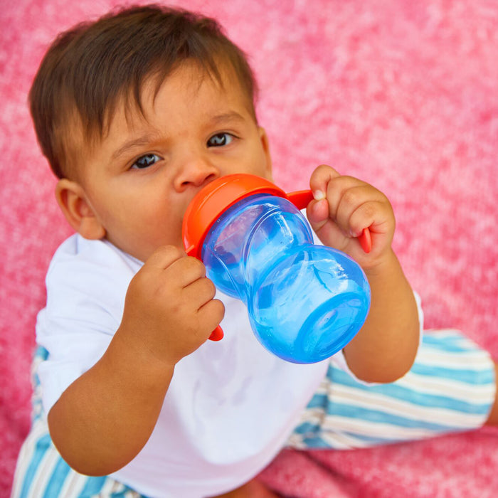 A baby in a white shirt and striped pants holds a Clik-It 2-Handle Sippy Cup (2 Pack) | Red / Yellow while sitting on a pink textured surface.