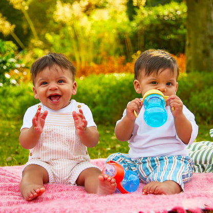 Two babies sit on a pink blanket outdoors. One baby claps and smiles, while the other holds a Clik-It 2-Handle Sippy Cup (2 Pack) | Red / Yellow and looks at the camera. Greenery and sunlight fill the background.