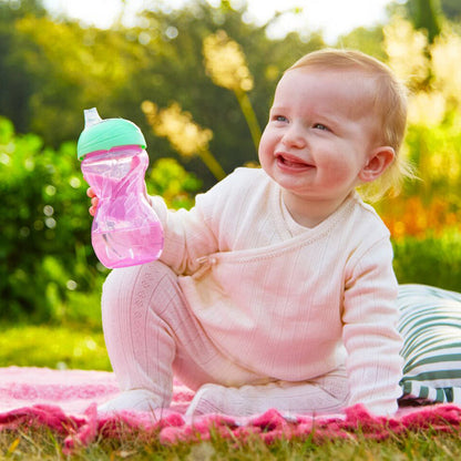 Smiling baby in a white outfit sits on a pink blanket outdoors, holding the Clik-It Soft Spout Sippy Cup (2 Pack) | Purple / Aqua. Tall grass and greenery can be seen in the background.