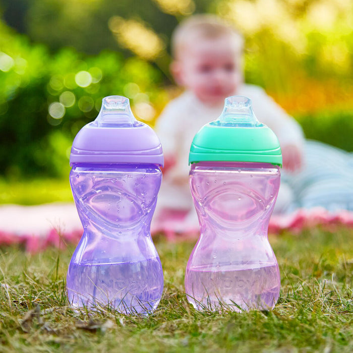 Two Clik-It Soft Spout Sippy Cup (2 Pack) | Purple / Aqua sit on grass outdoors, with a baby blurred in the background on a blanket. Both cups are spill-proof with soft spouts.