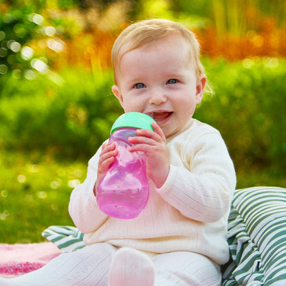 A smiling baby in white sits outdoors on a striped pillow, holding the Clik-It Soft Spout Sippy Cup (2 Pack) | Purple / Aqua. Blurred greenery and sunlight are in the background.