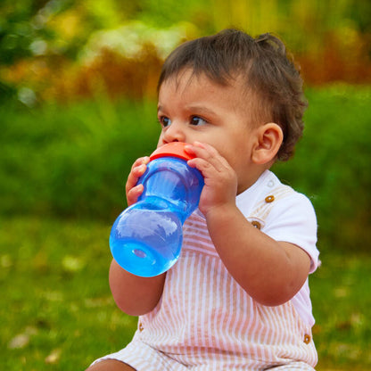 A toddler in a white outfit sits on grass, drinking from the Clik-It Soft Spout Sippy Cup (2 Pack) | Red / Yellow. Lush greenery and blurred plants create a gentle outdoor background.