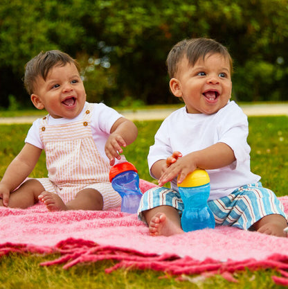 Two smiling babies sit on a pink blanket outdoors, each holding a Clik-It Soft Spout Sippy Cup (2 Pack) | Red / Yellow. They wear white shirts with striped overalls and shorts, surrounded by lush greenery.
