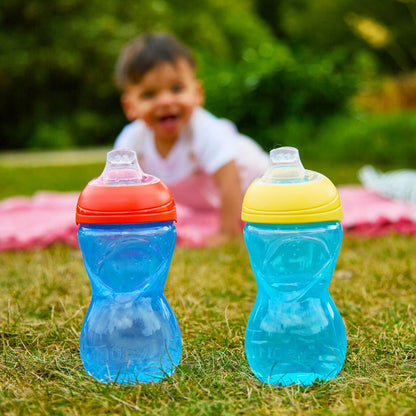 Two Clik-It Soft Spout Sippy Cup (2 Pack) | Red / Yellow sit on grass, one with a red lid and one with yellow. A toddler in a white shirt crawls on a pink blanket outdoors, blurred in the background.
