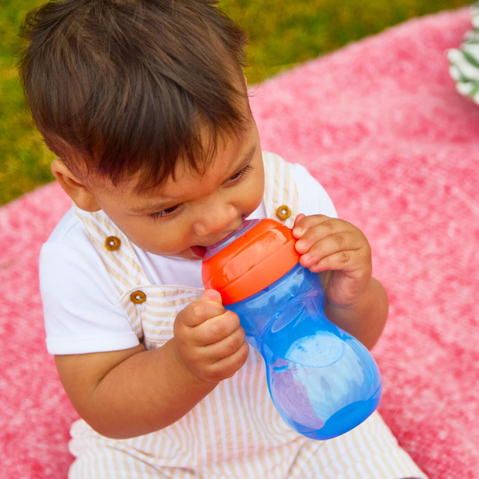 A toddler on a pink blanket enjoys a drink from the Clik-It Soft Spout Sippy Cup (2 Pack) | Red / Yellow, wearing a white shirt and striped overalls outdoors.