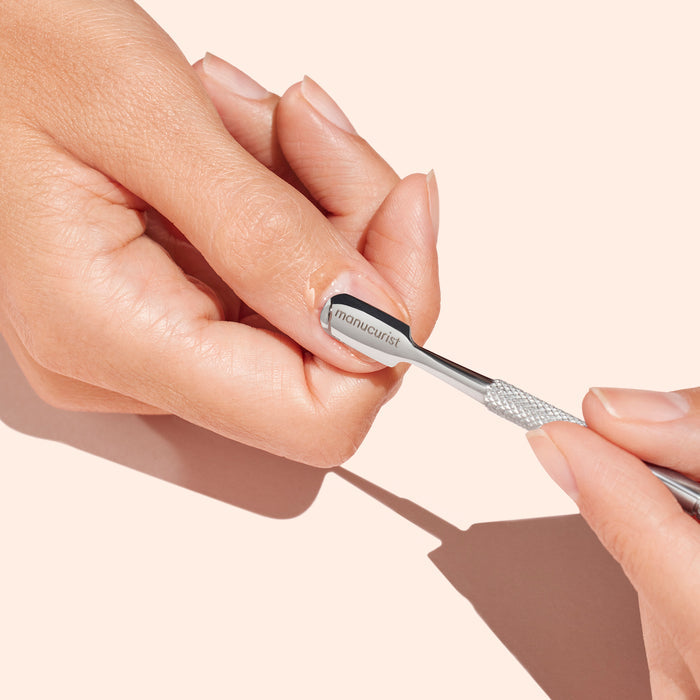 A person uses the Manucurist Cuticle Softener  Pusher Duo to gently push back cuticles and remove dead skin from the nail plate, set against a light peach background.