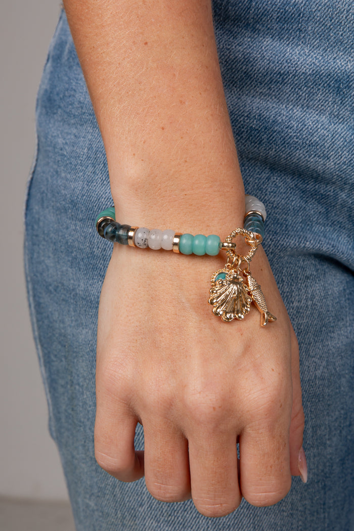 A close-up of a person's wrist wearing a beaded bracelet with turquoise, white, and dark gray beads, plus a gold-tone shell and starfish charm, against a blue denim background.