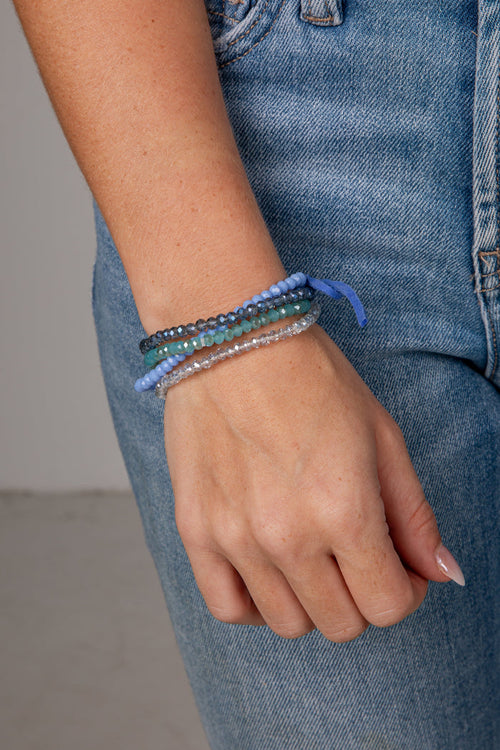 A close-up of a person's wrist wearing three beaded bracelets in shades of blue, teal, and clear, paired with blue denim jeans.