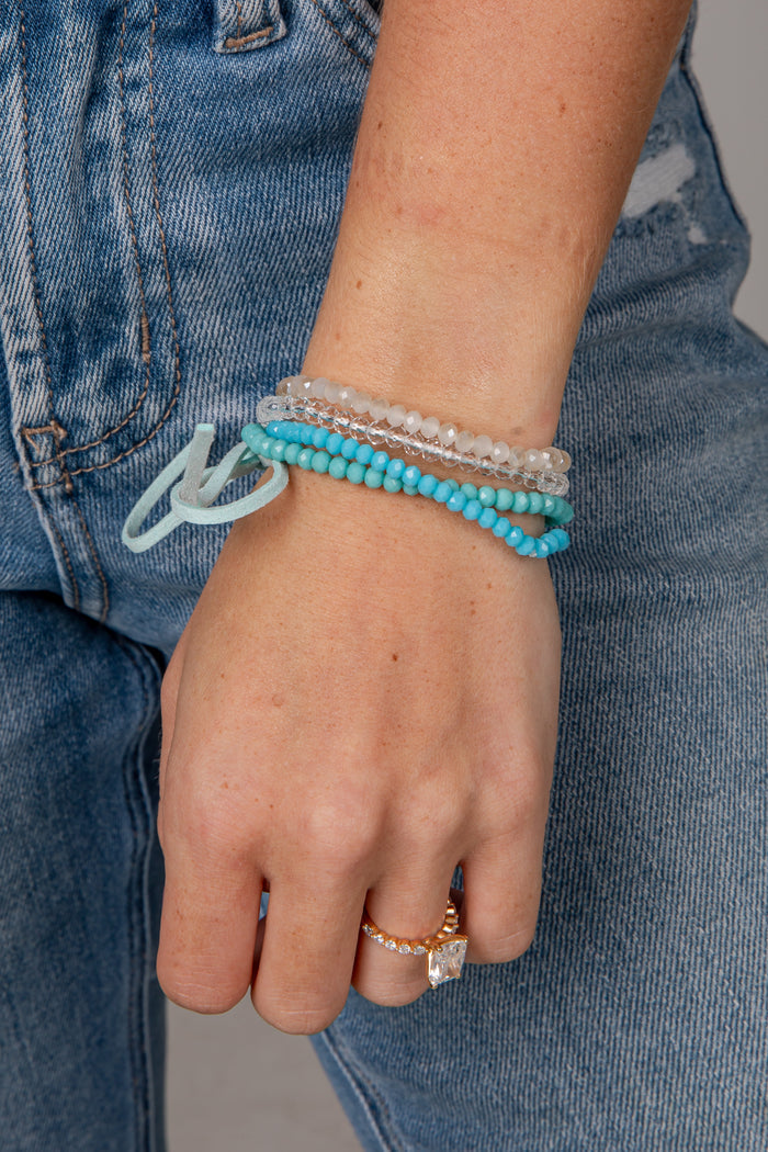 A close-up of a person's wrist wearing a stack of beaded bracelets, including turquoise, clear crystal, and light blue beads, paired with distressed blue jeans.