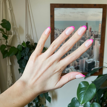 Flamingo French | Medium | Oval - Tab Press-On nails with neutral base and neon pink tips shown on hand indoors by plants and city art