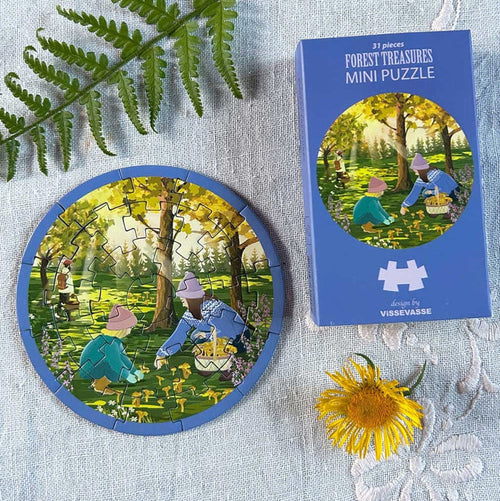 The Forest Treasure | 31 Piece Jigsaw Puzzle by ViSSEVASSE and its box feature an enchanting illustration of people gathering mushrooms in a sun-dappled forest. Nearby, a yellow flower and green fern rest on light-colored fabric, inviting you to creatively immerse yourself as you piece together this tranquil scene.