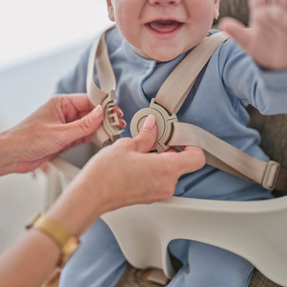 An adult secures a smiling baby in a Grow with Me Foldable High Chair by fastening the safety harness. The baby, dressed in a blue outfit, raises one hand as the adult adjusts the beige straps.