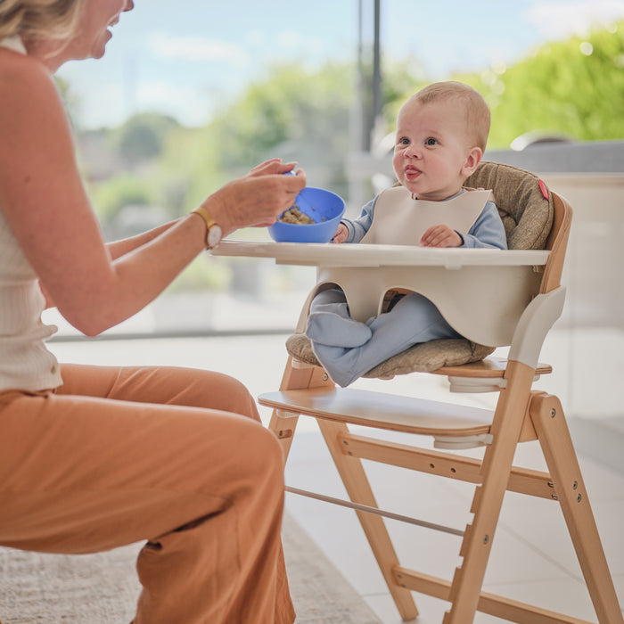 A woman feeds a baby sitting in a Grow with Me Foldable High Chair. The baby, wearing a bib, gazes attentively at the spoon as bright natural light fills the modern, airy room.