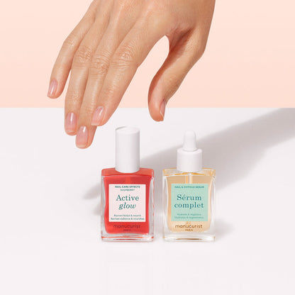 A hand with neatly manicured nails hovers above the Manucurist Healthy Glow Duo—two nail care bottles—on a white surface against a soft pink background.