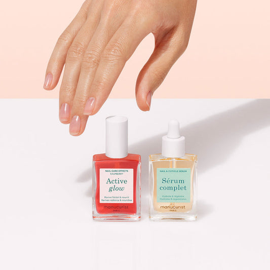 A hand with neatly manicured nails hovers above the Manucurist Healthy Glow Duo—two nail care bottles—on a white surface against a soft pink background.