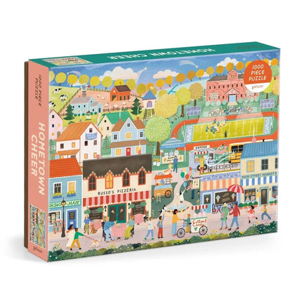 The Galison Hometown Cheer 1,000 Piece Jigsaw Puzzle features an illustrated small town scene with shops, houses, people, and trees on the box.