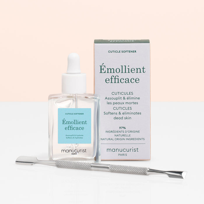 A bottle of Manucurist Hydrating Cuticle Softener with a dropper, its branded box, and a metal cuticle pusher tool are arranged on a light surface with a soft pink background—ideal for achieving an at-home manicure.