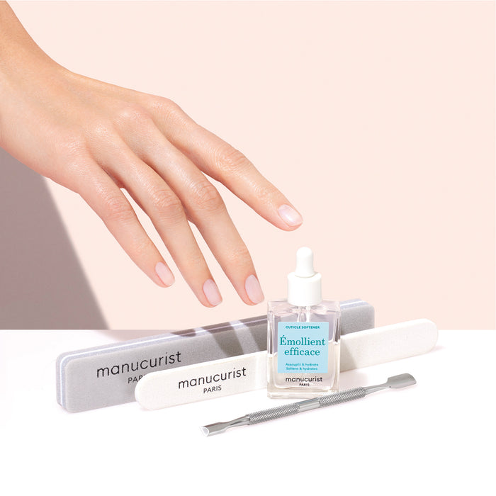 A hand with manicured nails hovers over Manucurist nail care essentials, including the Hydrating Cuticle Softener, emollient oil, nail files, and a cuticle pusher—all set against a soft pink background.