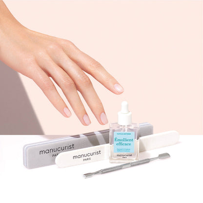 A hand with manicured nails hovers over Manucurist nail care essentials, including the Hydrating Cuticle Softener, emollient oil, nail files, and a cuticle pusher—all set against a soft pink background.