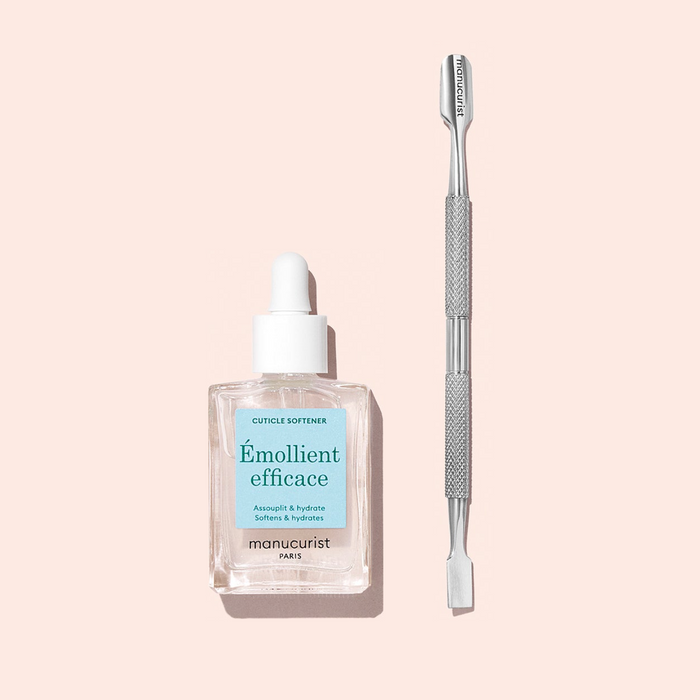 A bottle of Manucurist Hydrating Cuticle Softener, complete with a convenient dropper, sits alongside a metal cuticle pusher on a light pink background—creating the perfect duo for achieving a flawless manicure.