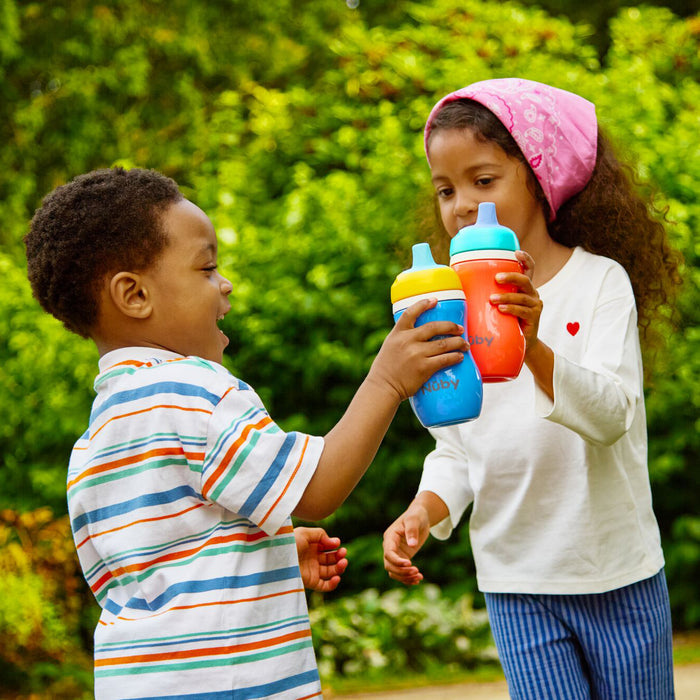 A smiling boy in a striped shirt and a girl in a pink headscarf clink Insulated Active Sipper Cup (2 Pack) outdoors, surrounded by green foliage.