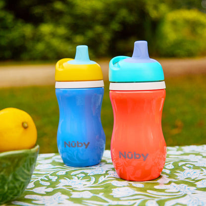 Two leak-resistant Insulated Active Sipper Cup (2 Pack) sippy cups, one blue/yellow and one red/blue, sit on a floral tablecloth outside, with a bowl of lemons to the left and greenery in the background.