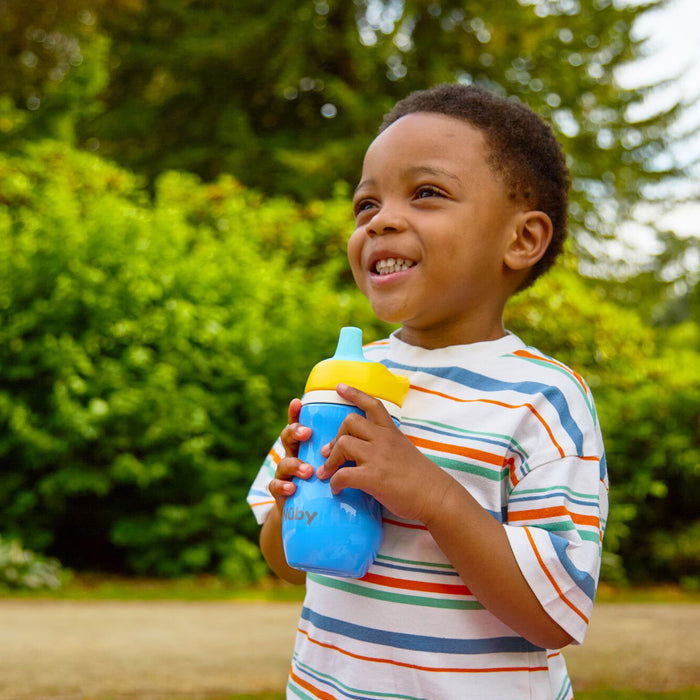 Smiling young boy in a striped shirt holds an Insulated Active Sipper Cup (2 Pack) with a hard spout and yellow lid while standing outdoors in front of green bushes.