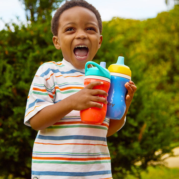A young boy in a striped shirt stands outside, smiling with his mouth open as he holds a red and a blue Insulated Active Sipper Cup (2 Pack), with green bushes and trees in the background.