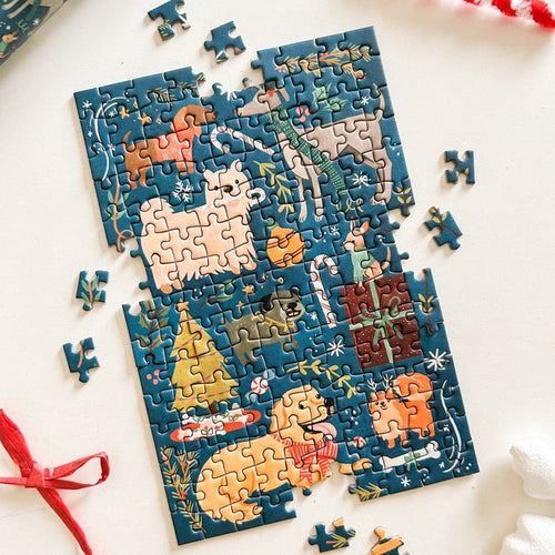 The partially completed Merry Woofmas | 150 Piece Jigsaw Puzzle by Spaghetti Tree features adorable animal illustrations, a Christmas tree, and festive decorations on a white surface. Loose pieces from this charming portable puzzle are scattered around, making it perfect for cozy holiday moments anywhere.