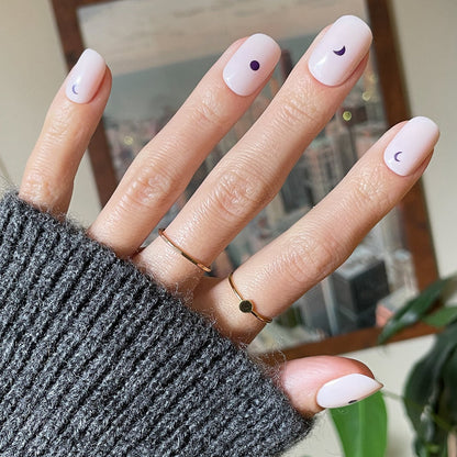 Moon Phases | Short | Squoval - Tab Press-On nails with pale base and small dark moon phase designs on hand indoors