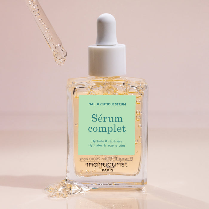 A dropper releases Manucurist Nail Hydrating Serum into a clear glass bottle labeled with the brand name. The soft beige background highlights droplets of this nourishing nail treatment visible on the bottle.