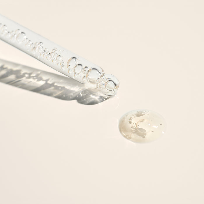 A clear glass dropper with air bubbles and a drop of Manucurist Nail Hydrating Serum rest on a light beige surface, casting a soft shadow—ideal for nail treatments or repairing damaged nails.